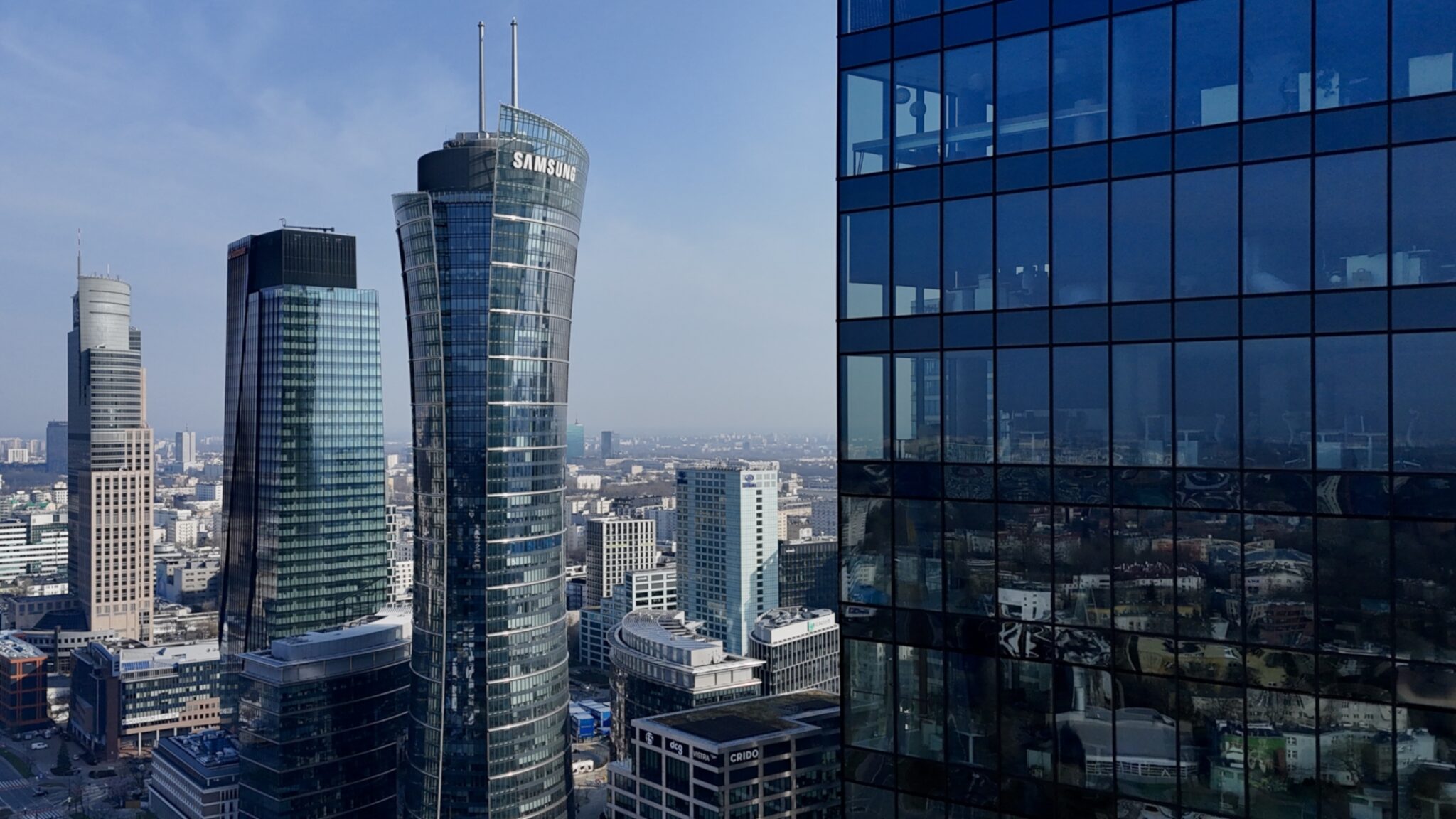 Centrum biznesowe z obłym szklanym wieżowcem oraz ciemną elewacją biurowca odbijającą zabudowę miejską