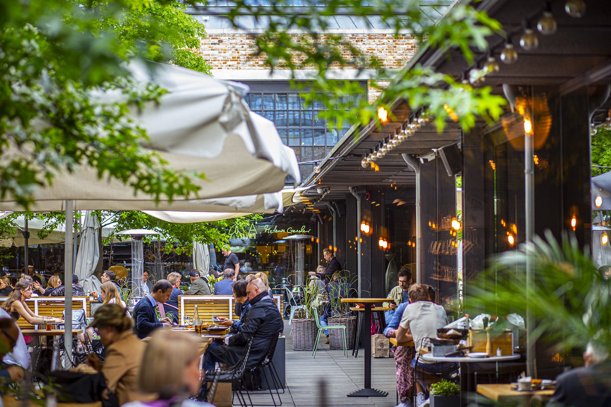 Zewnętrzny ogródek restauracyjny wypełniony gośćmi pod parasolami w otoczeniu industrialnej cegły i zielonych roślin.
