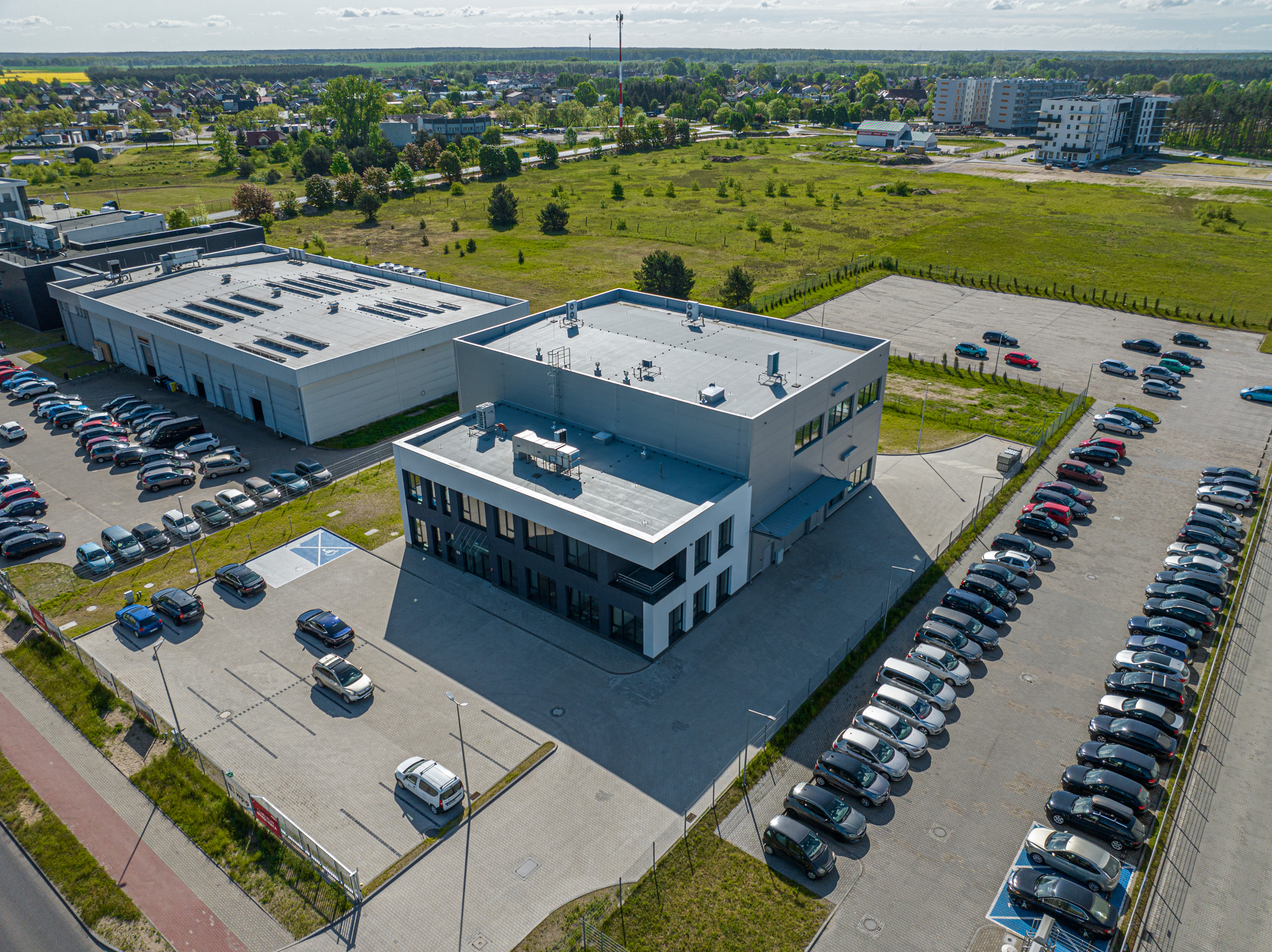 Widok z lotu ptaka na kompleks biurowo-magazynowy z rozległymi parkingami samochodowymi na tle podmiejskiego krajobrazu.