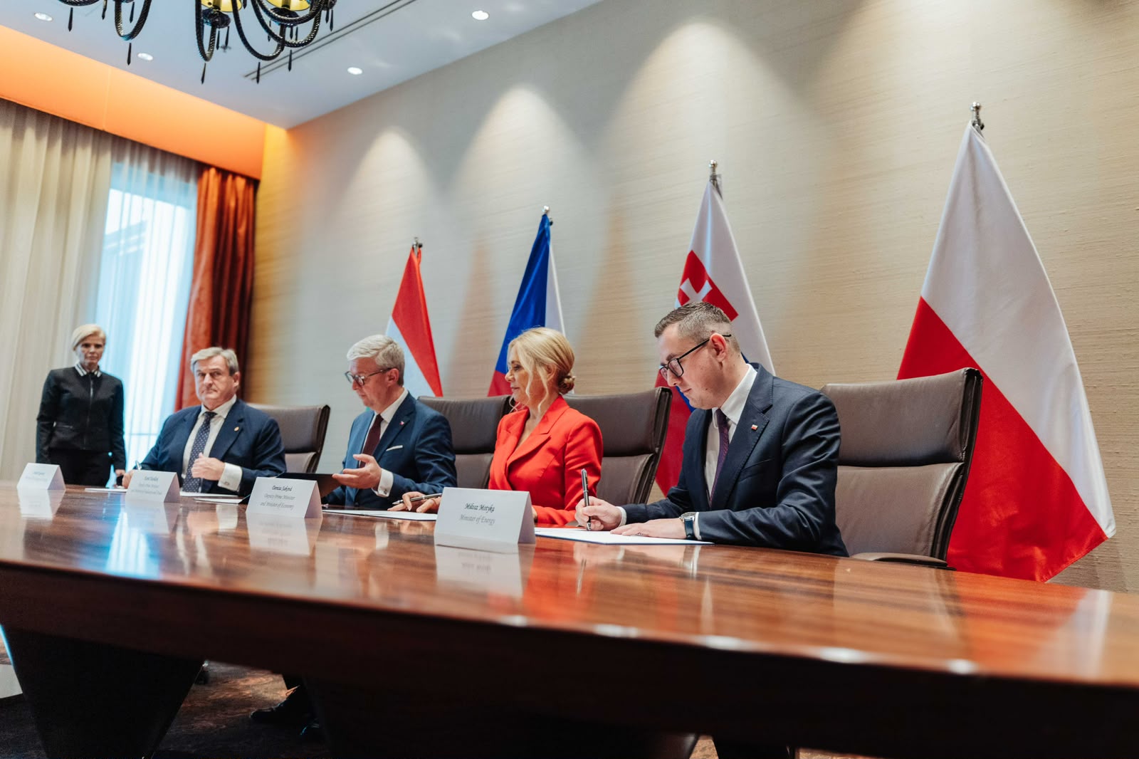 Przedstawiciele państw Grupy Wyszehradzkiej siedzą przy stole konferencyjnym na tle flag państwowych i podpisują dokumenty.