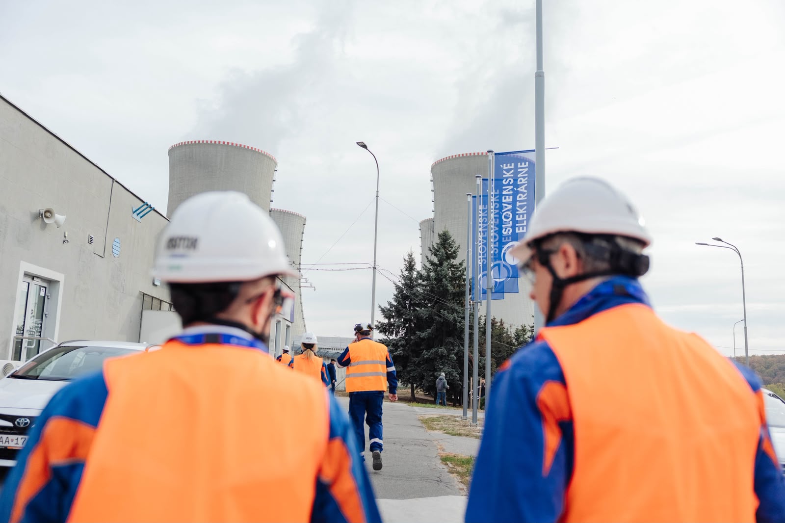 Pracownicy w kaskach i odblaskowych kamizelkach idą drogą w stronę chłodni kominowych elektrowni.