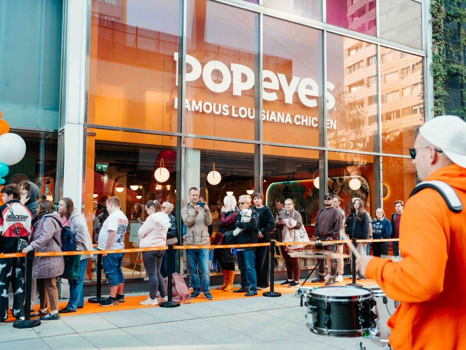 Długa kolejka klientów przed przeszkloną witryną restauracji Popeyes oraz bębniarz w pomarańczowej bluzie podczas otwarcia lokalu.