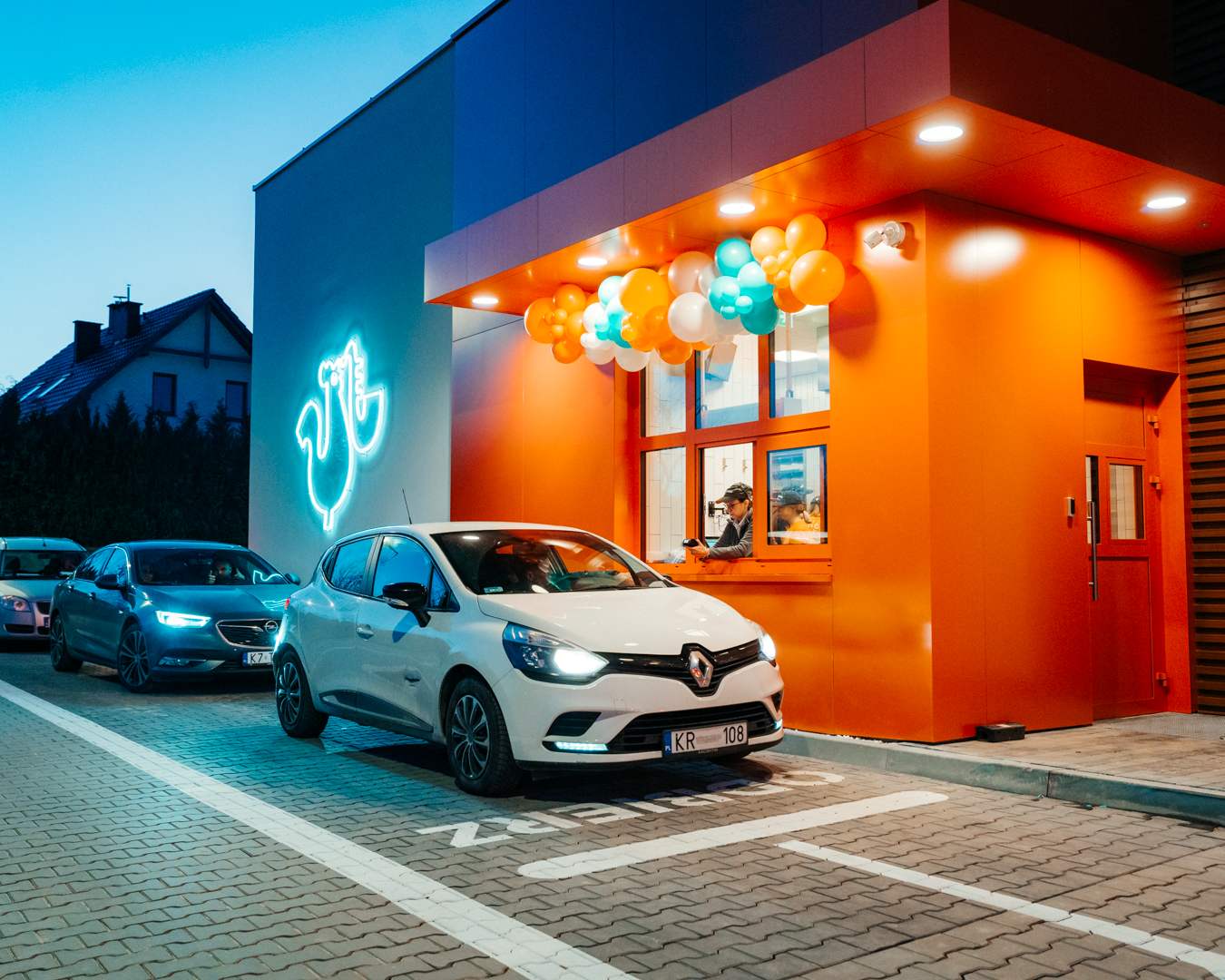 Widok zewnętrzny restauracji Popeyes o pomarańczowej elewacji z okienkiem drive-thru i oczekującymi samochodami o zmierzchu.