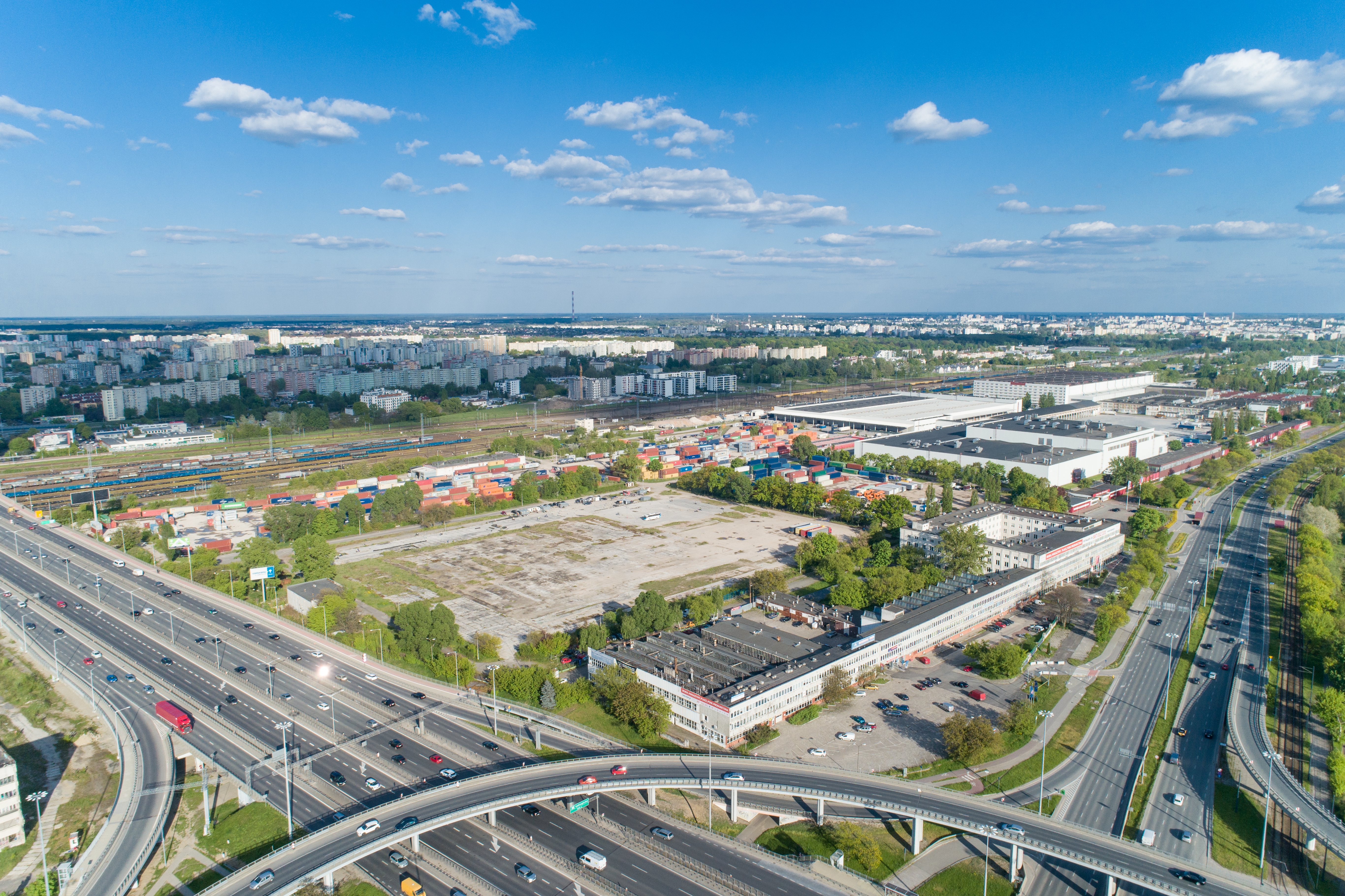 Widok z góry na węzeł drogowy, halę magazynową, place i biurowce na tle panoramy miejskiej.