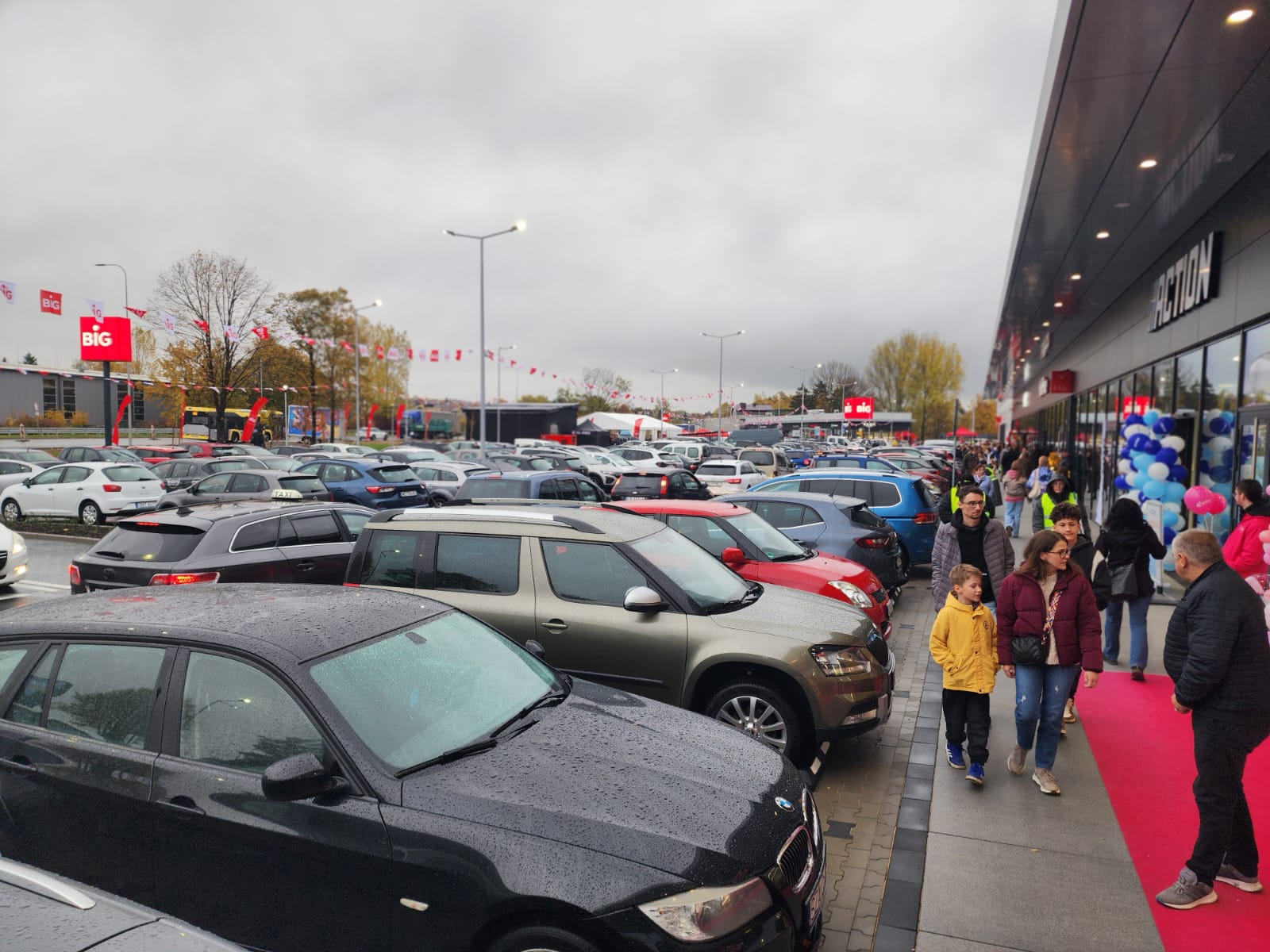 Zatłoczony parking i klienci spacerujący wzdłuż witryn sklepów nowego parku handlowego BIG w deszczowy dzień.