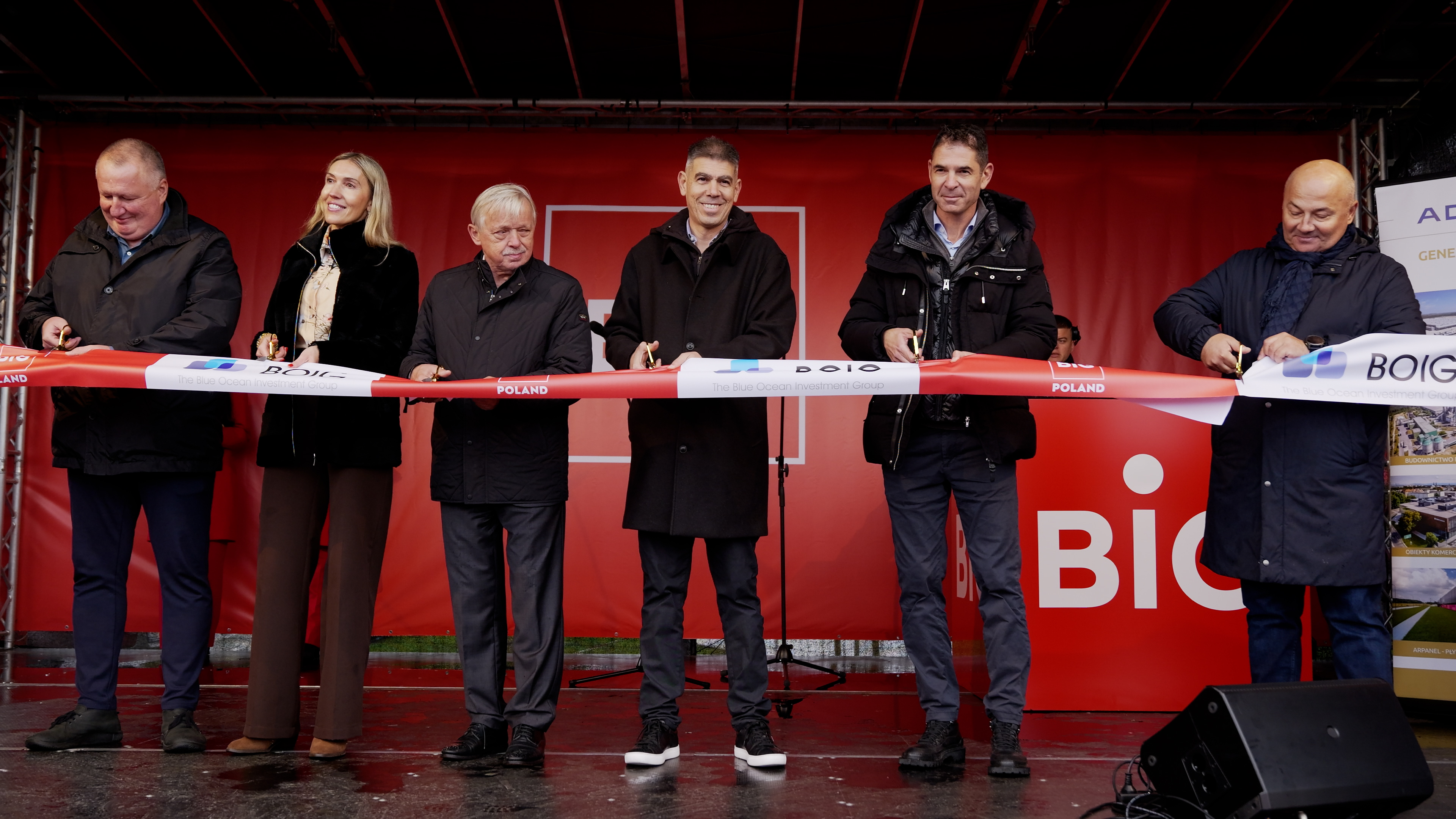 Sześć osób przecina biało-czerwoną wstęgę na scenie z czerwonym tłem BIG Poland podczas uroczystego otwarcia obiektu.