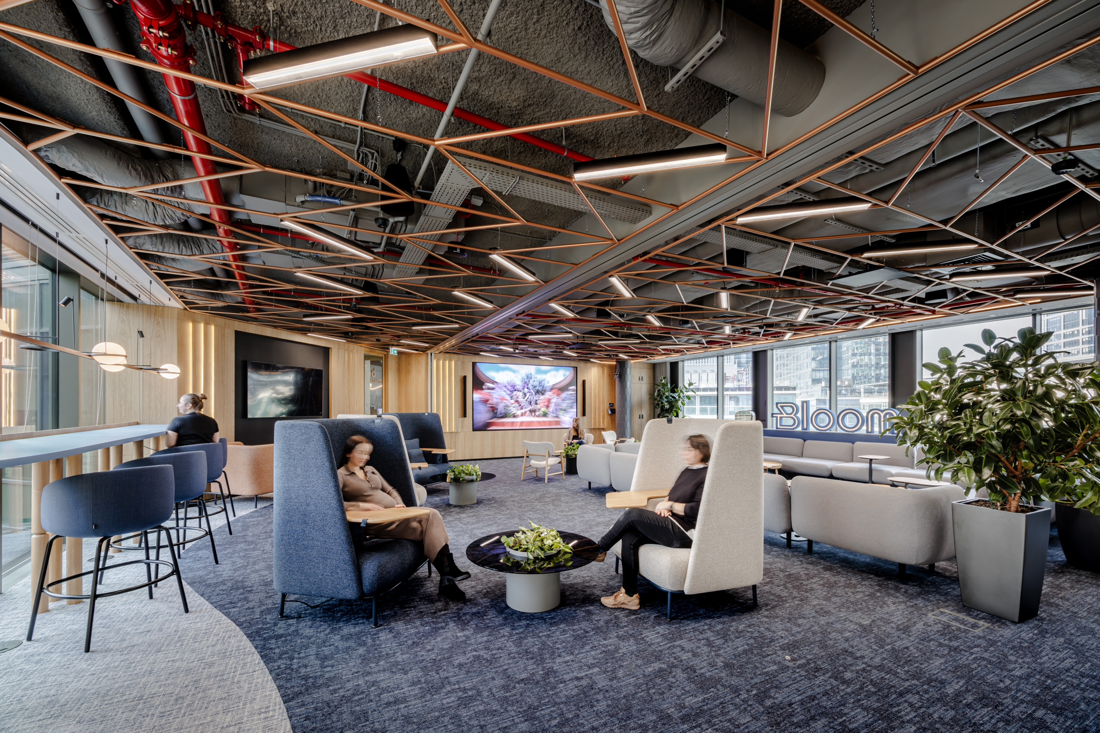 Nowoczesna przestrzeń biurowa lounge z miedzianą konstrukcją sufitu, dźwiękochłonnymi fotelami i przeszkleniami z widokiem na panoramę miasta.