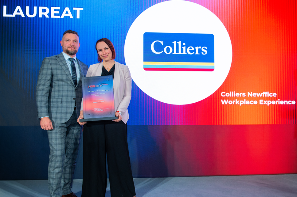 Uśmiechnięta kobieta i mężczyzna stojący na scenie z tablicą pamiątkową na tle wielkoformatowego ekranu z logo firmy Colliers.