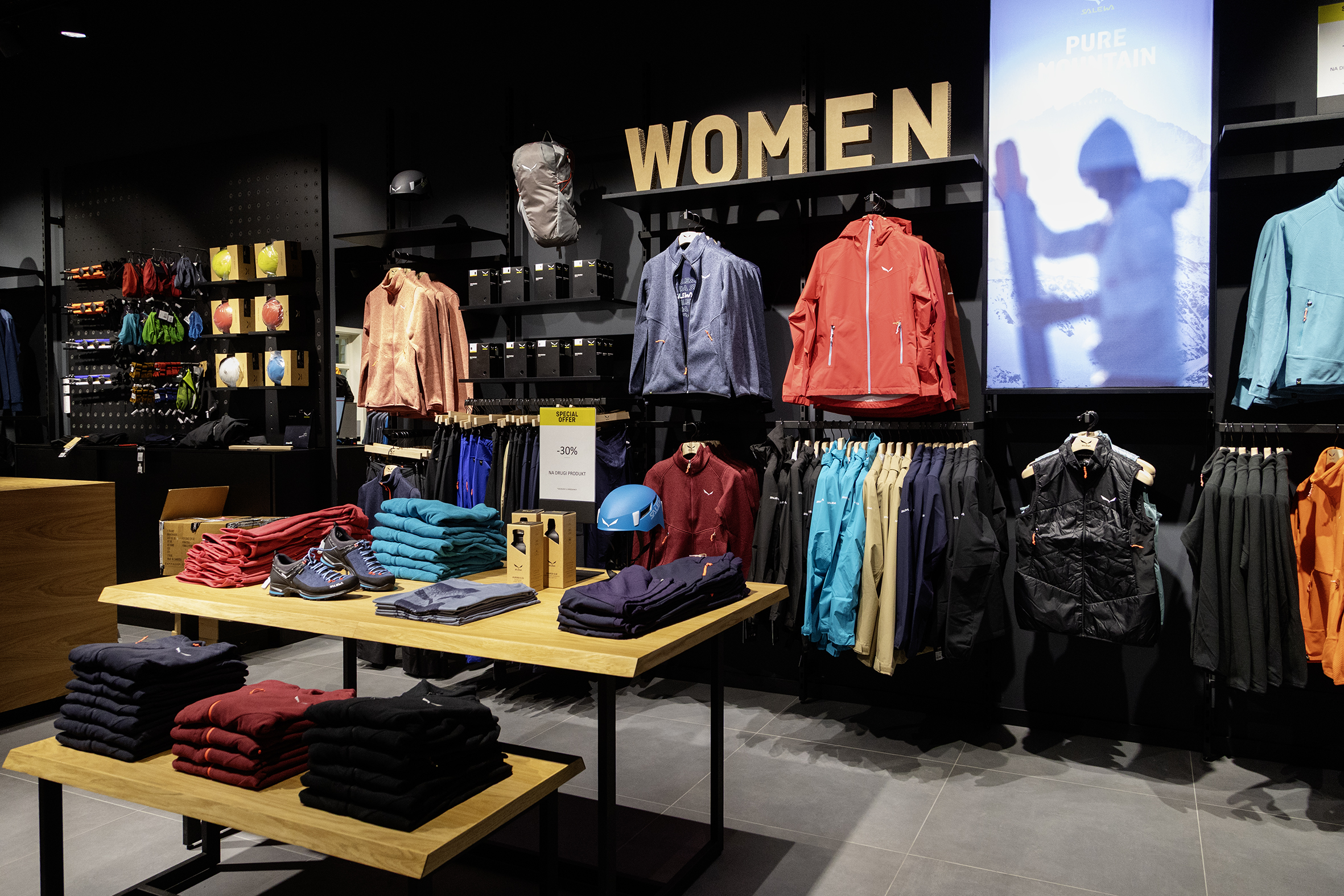 Nowoczesne wnętrze sklepu z sekcją damską WOMEN, kolorową odzieżą górską, butami trekkingowymi i kaskiem na drewnianym stole.