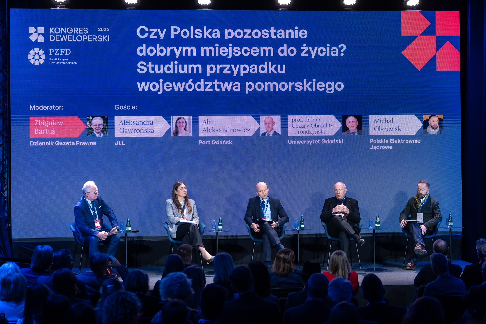 Pięciu panelistów w strojach biznesowych dyskutuje na scenie podczas Kongresu Deweloperskiego na tle multimedialnego ekranu.