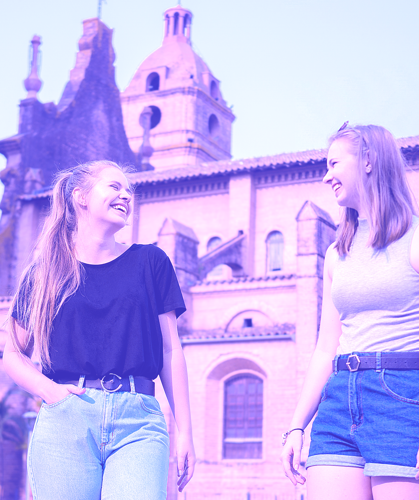 Twee jonge vrouwen die glimlachen en buiten wandelen met historische stenen gebouwen op de achtergrond.