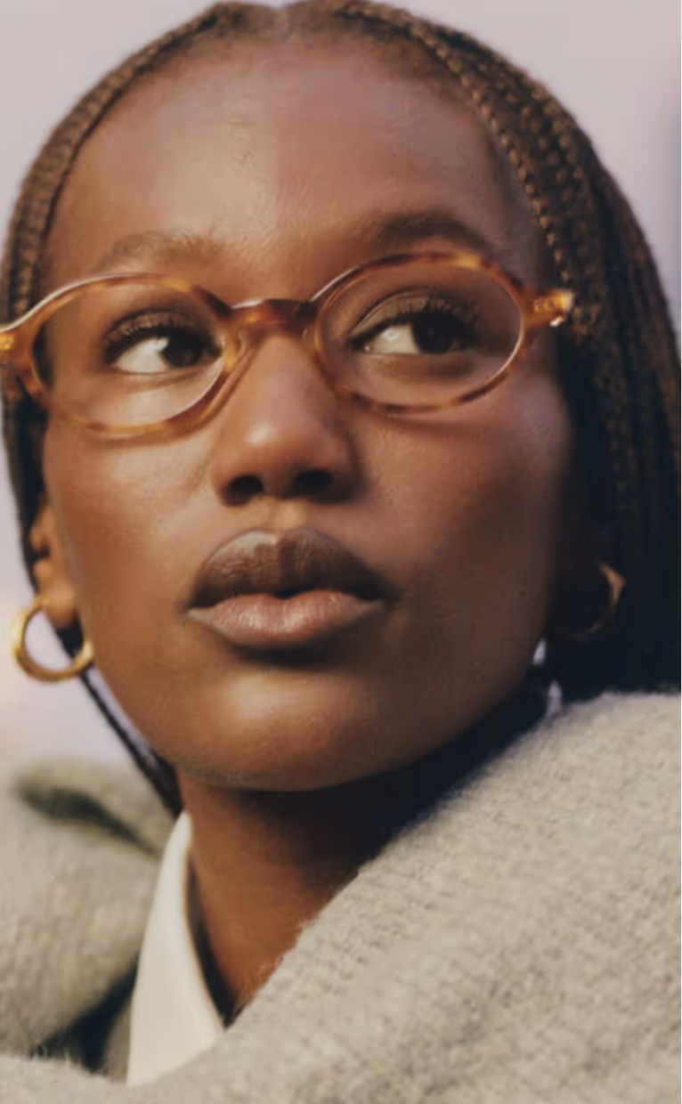 Close-up of a thoughtful Black woman with braided hair wearing round tortoiseshell glasses, gold hoop earrings, and a grey sweater.