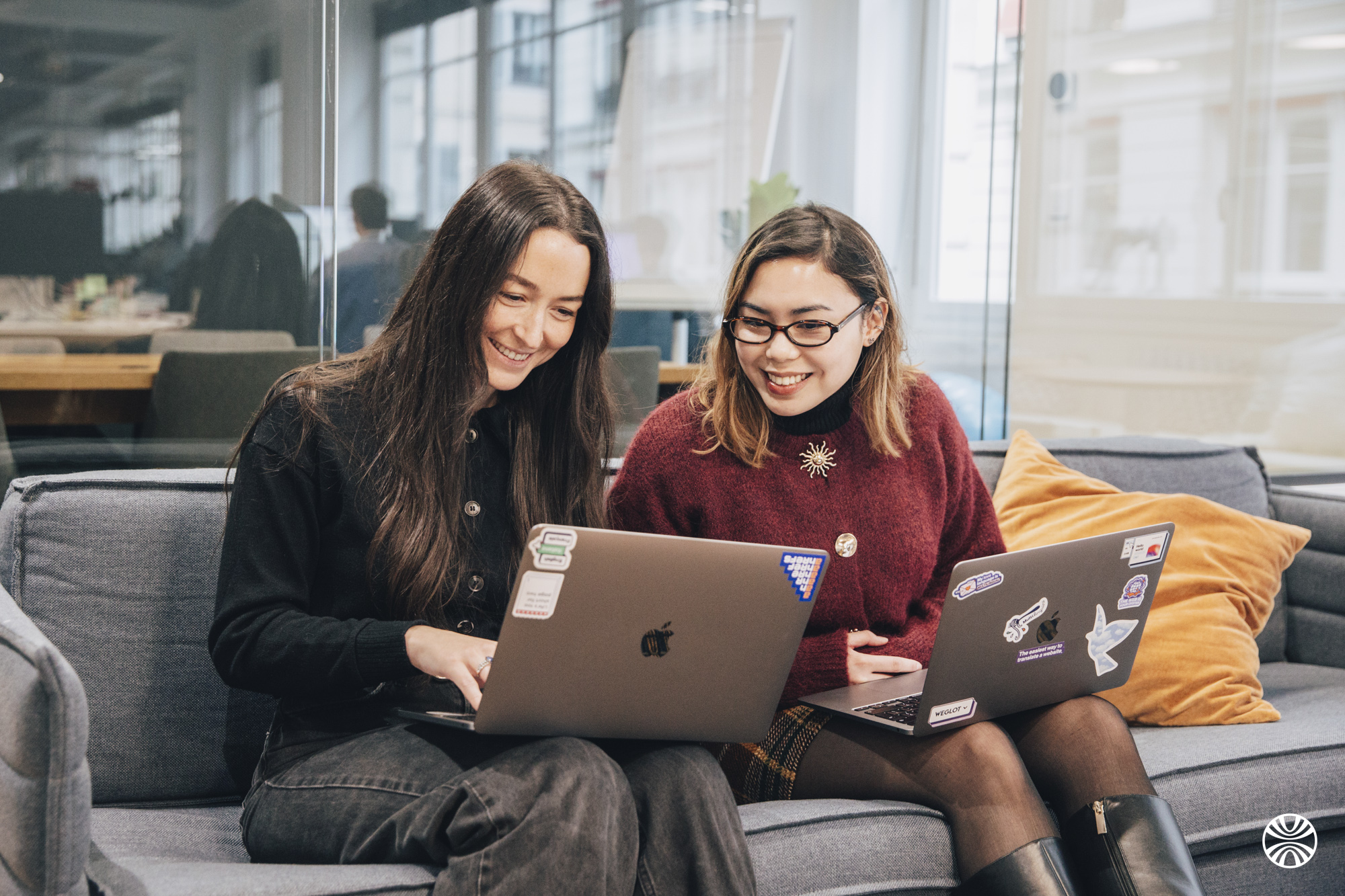 Twee vrouwen zitten op een grijze bank, glimlachen en kijken naar laptops met stickers in een moderne kantooromgeving.