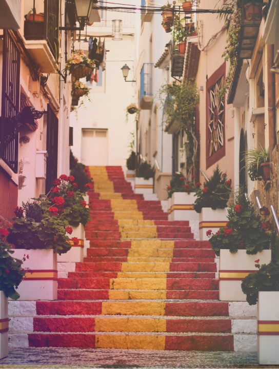 Outdoor stairway decorated with red and yellow paint and lined with flower pots along a narrow street with hanging plants and lanterns.