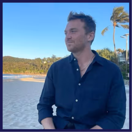 Man with tousled hair wearing a dark shirt sitting on a sandy beach with a palm tree in the background.