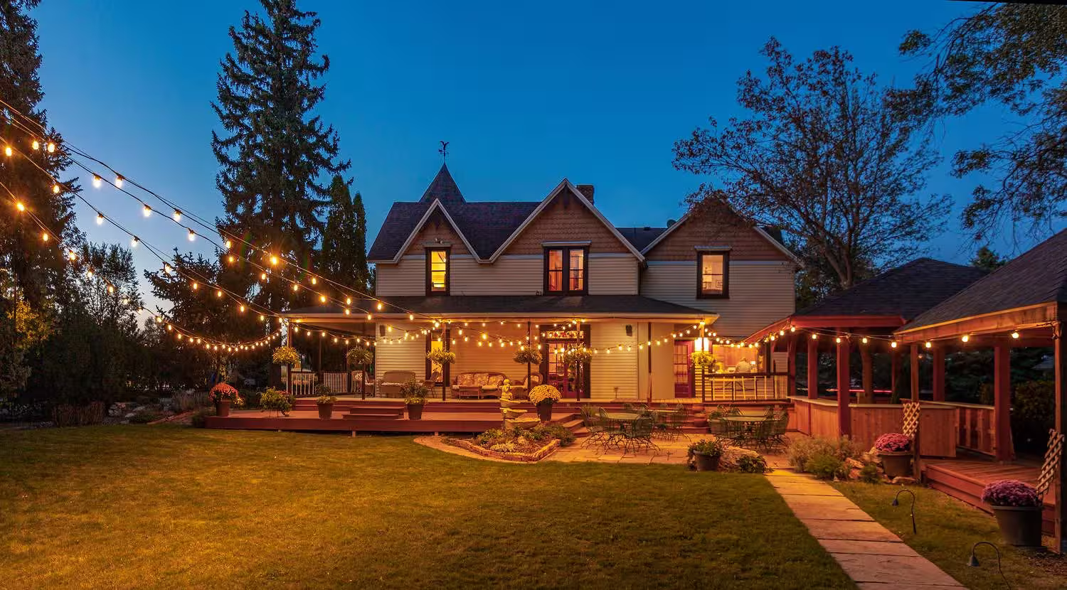 Two-story house with a porch decorated with string lights and surrounding garden at dusk.