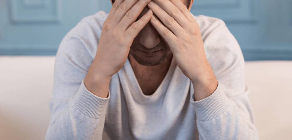 Young man wearing a light gray sweater sitting with his head in his hands, appearing stressed or sad.