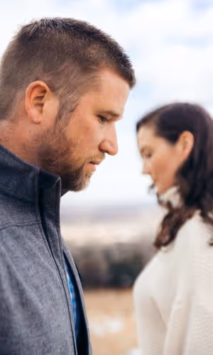 Close-up side profile of a man with a beard looking down, with a blurred woman in white in the background outdoors.