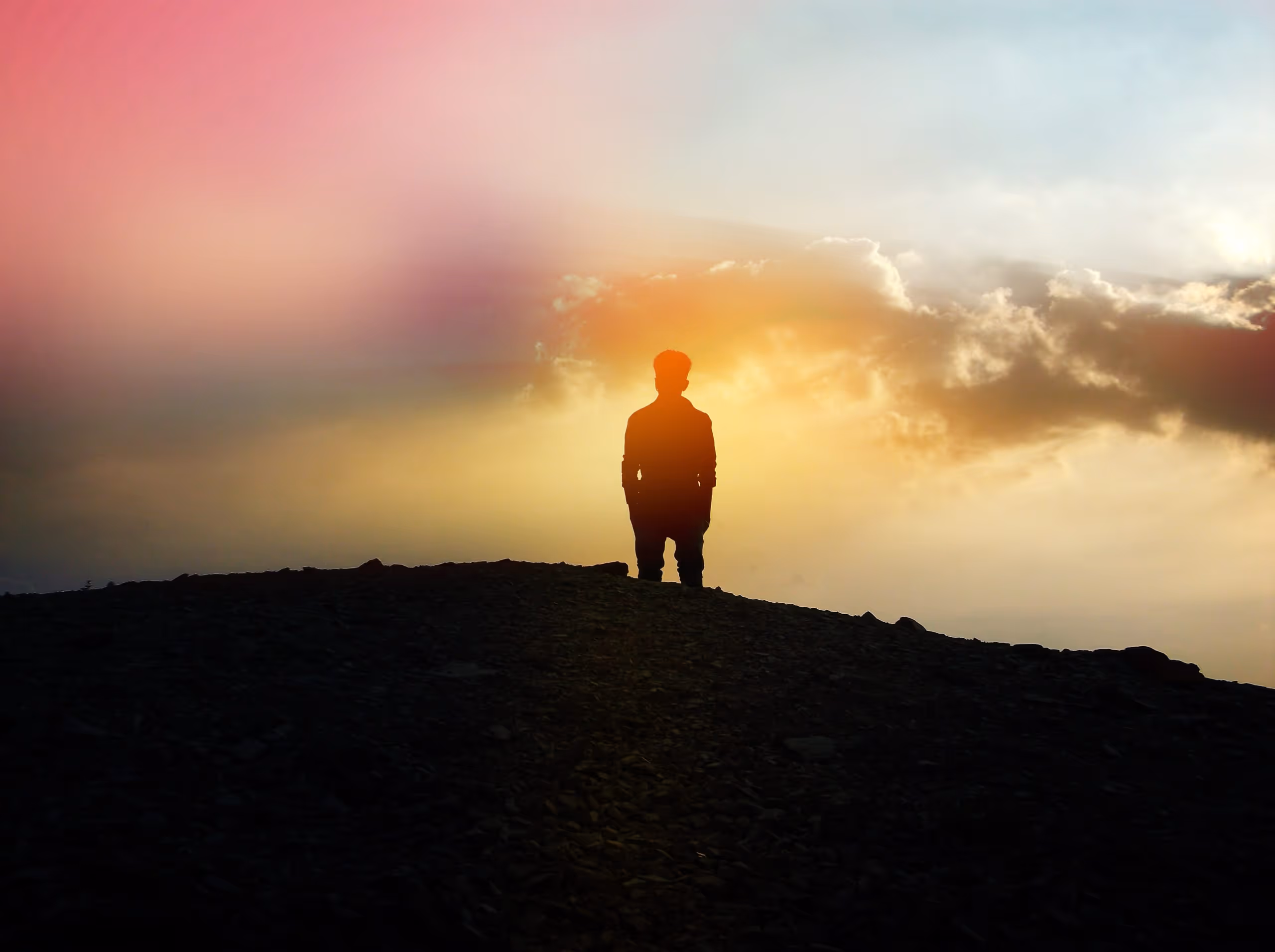 Silhouette of a person standing on a hill during a colorful sunset with clouds.