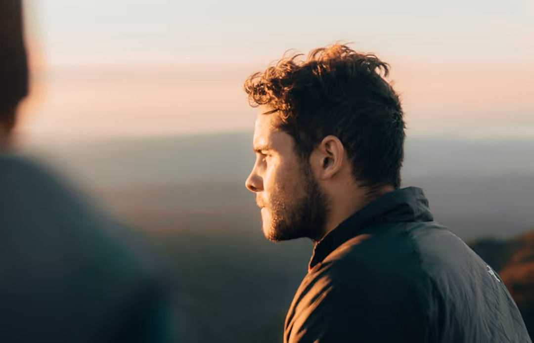 Side profile of a young man with curly hair and a beard, looking thoughtfully into the distance during sunset.