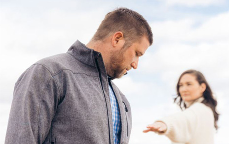 Man in gray jacket looking downward with a woman in white reaching out toward him in the background under a cloudy sky.