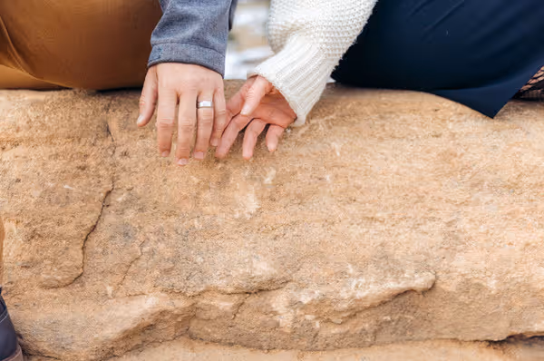 A couple holding hands over a textured rock surface, showing their fingers intertwined with one person wearing a silver ring.