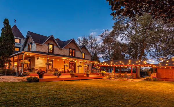 Large house with warm exterior lights, decorated porch, and string lights over a patio at dusk.