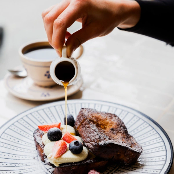 Social media content creation - Hand pouring syrup onto French toast topped with cream, blueberries, and strawberry slices, with a cup of coffee in the background.