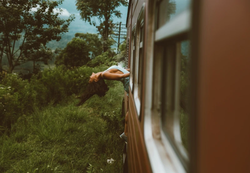 A woman leans out of a moving train window with her head tilted back and hair flowing, surrounded by green trees and grass.