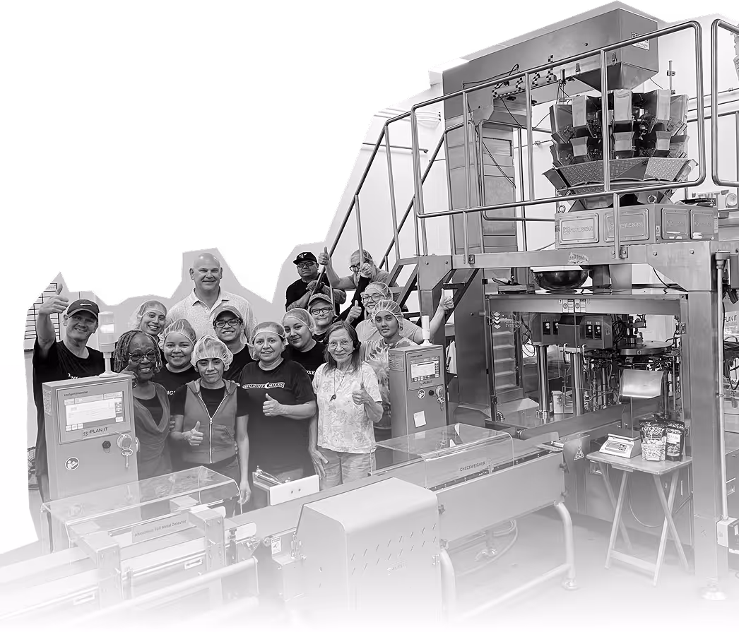 Group of factory workers wearing hairnets standing together and giving thumbs up next to food packaging machinery.