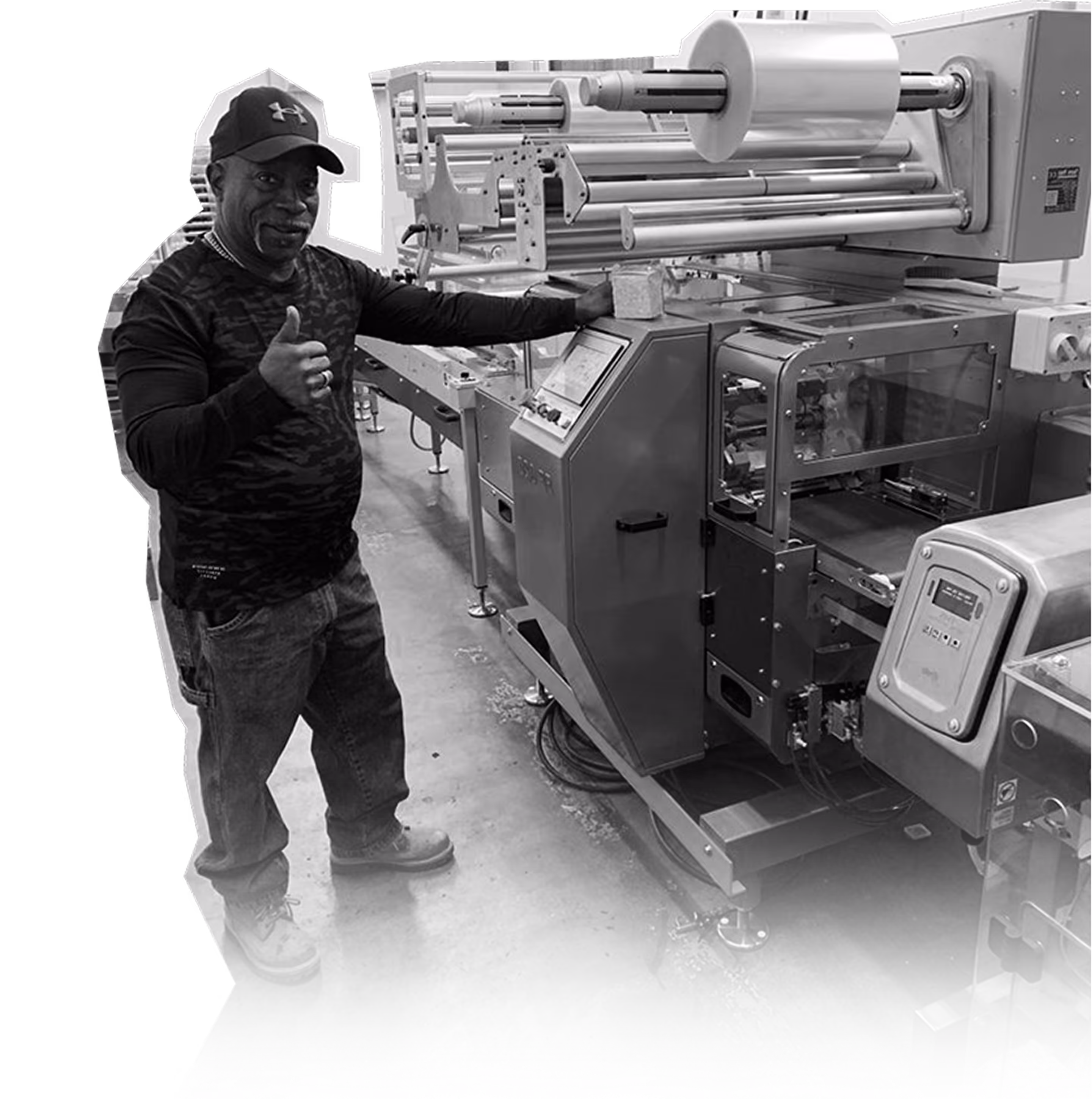 Man wearing a cap and camouflage long sleeve shirt giving a thumbs-up next to industrial packaging machinery.