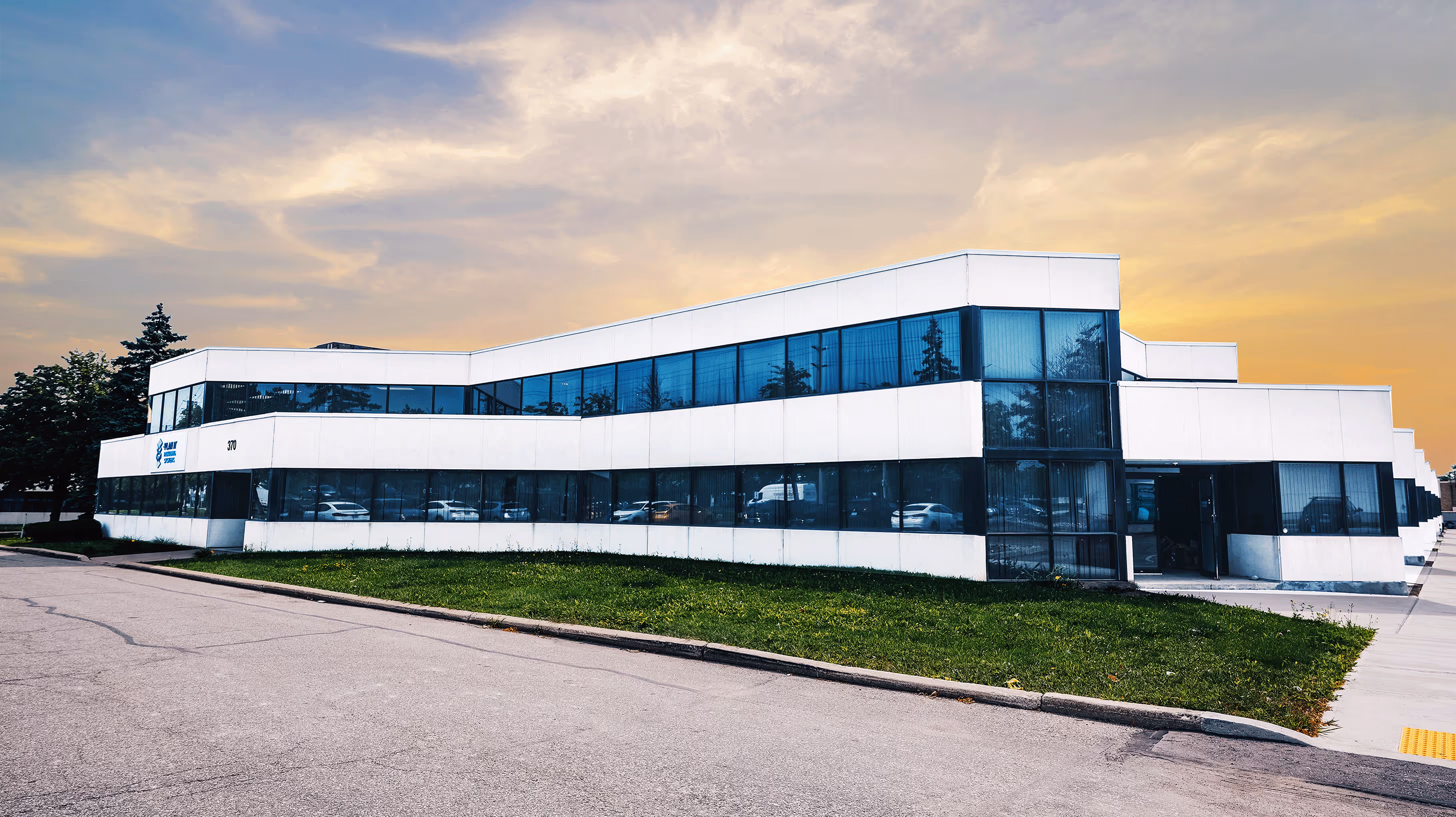 Modern two-story white office building with large blue-tinted windows and a green lawn under a cloudy sunset sky.