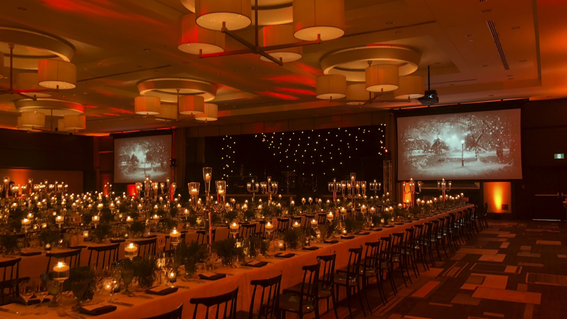 Elegant corporate holiday party with warm amber event lighting, starry sky stage backdrop, candlelit tables, and dual projection screens displaying vintage winter scenes. AV production by Contak AV in Montreal.