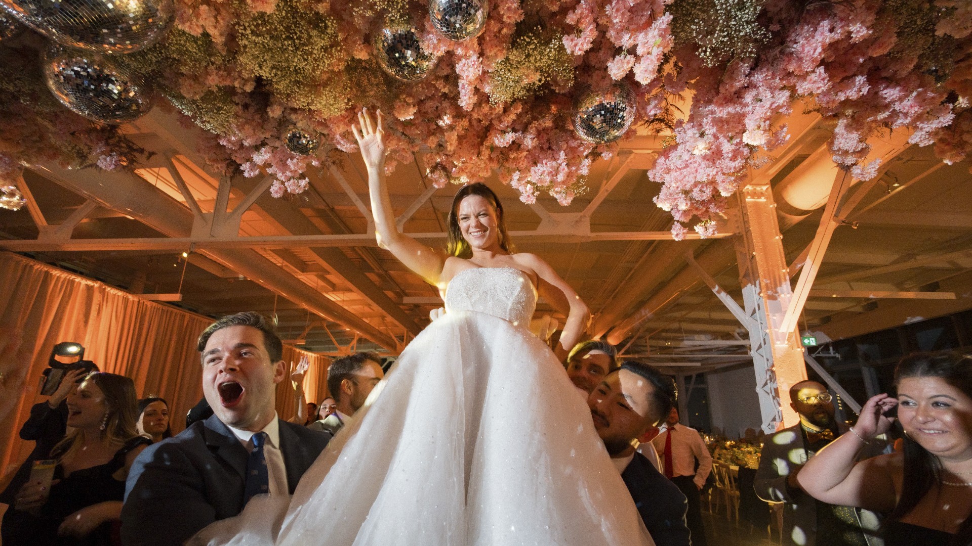 High-energy wedding moment with bride raised on shoulders, custom lighting design, hanging floral disco decor, and soft ambient uplighting
