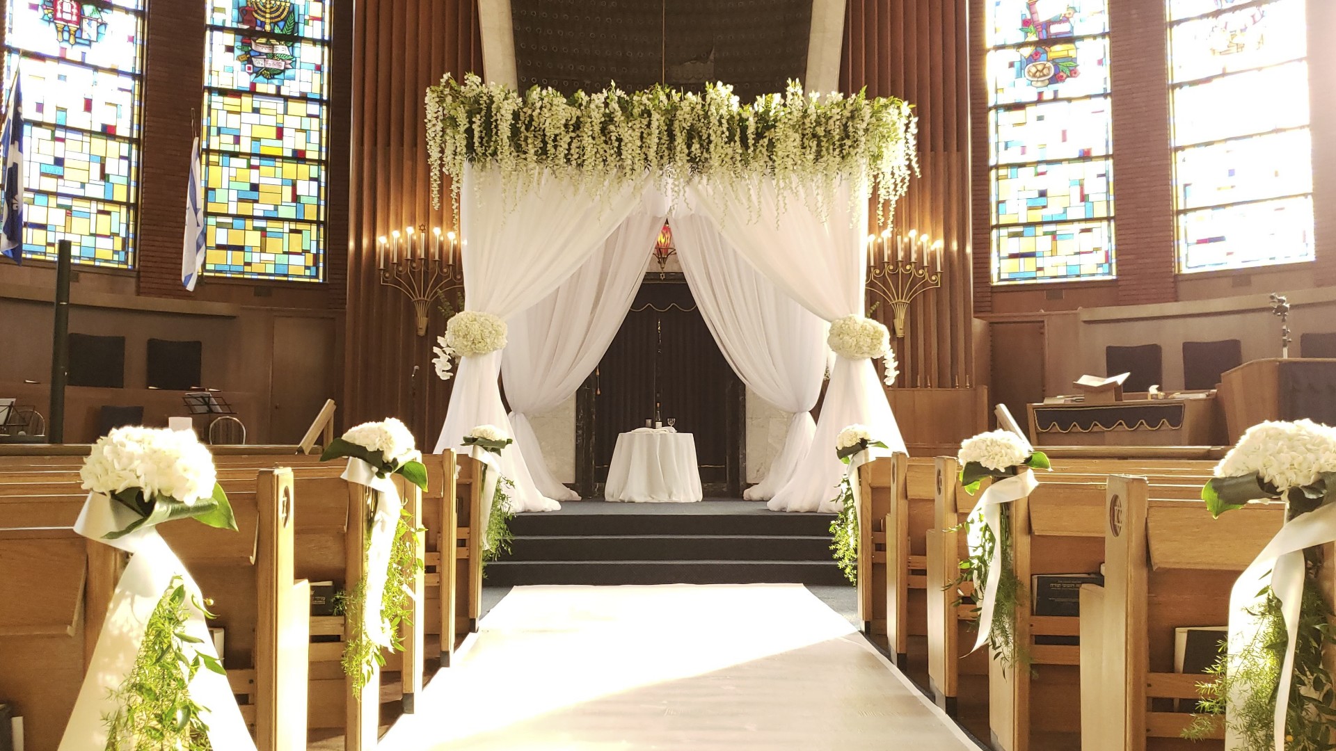 White Chuppah installation with sheer draping and white floral aisle arrangements at Shaar Hashomayim synagogue in Montreal
