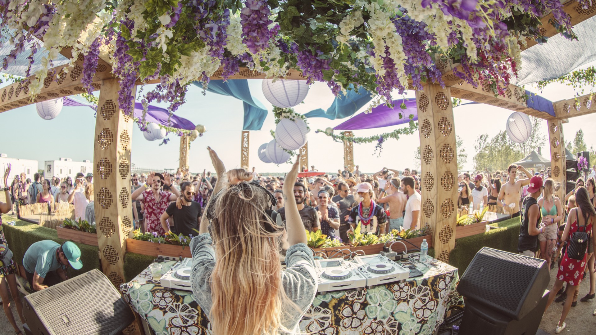 Sunlit Montreal music festival with vibrant overhead decor, custom wooden DJ booth, and a female DJ energizing the crowd with hands in the air