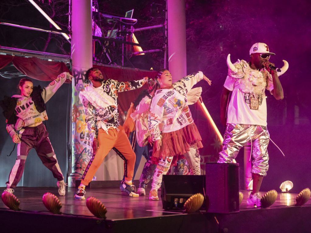Synchronized breakdancers performing under dramatic stage lighting and clamshell footlights as rapper delivers live performance at Montreal cultural event