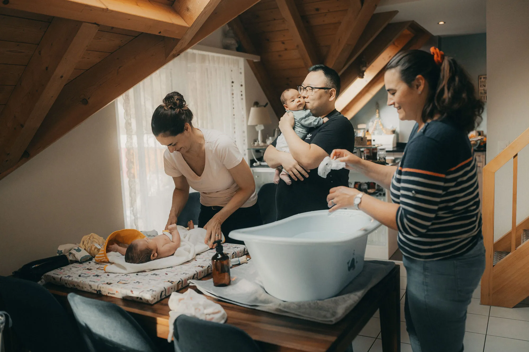 Vater hält ein Baby und küsst es, Mutter wickelt das zweite Baby und Hebamme steht neben dran mit Badewanne