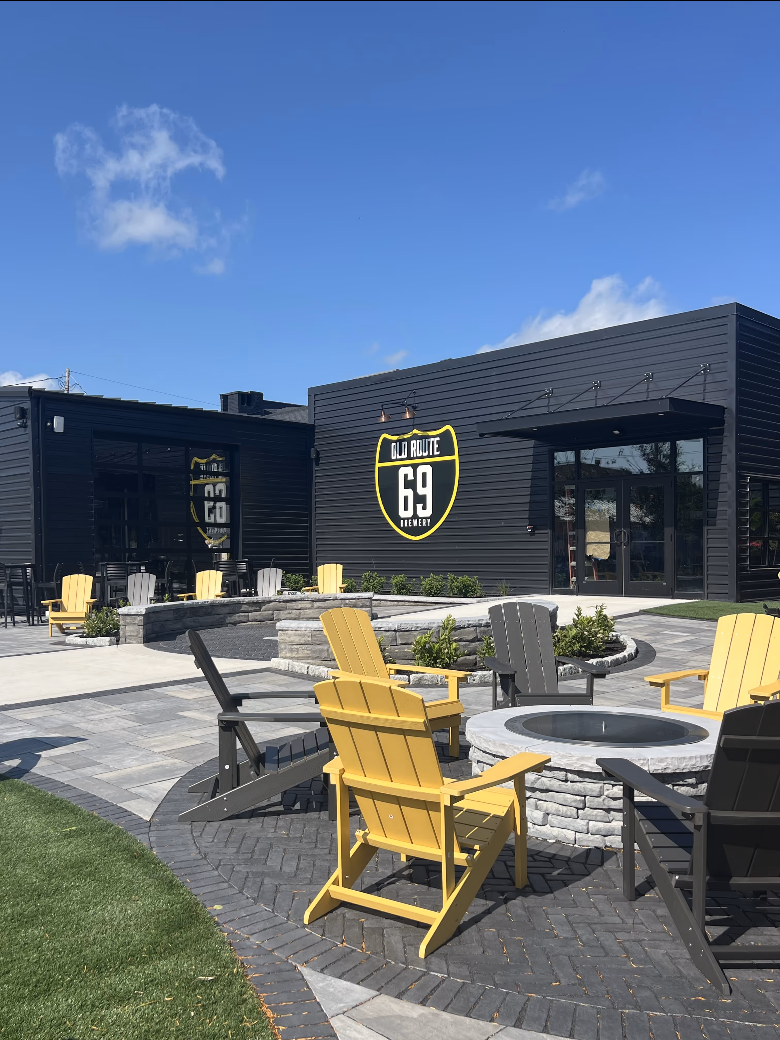 Exterior of Old Route 69 Brewery in Ada, Ohio, showcasing outdoor seating with Adirondack chairs arranged around a stone fire pit, set against the backdrop of the brewery’s modern building and landscaped lawn.