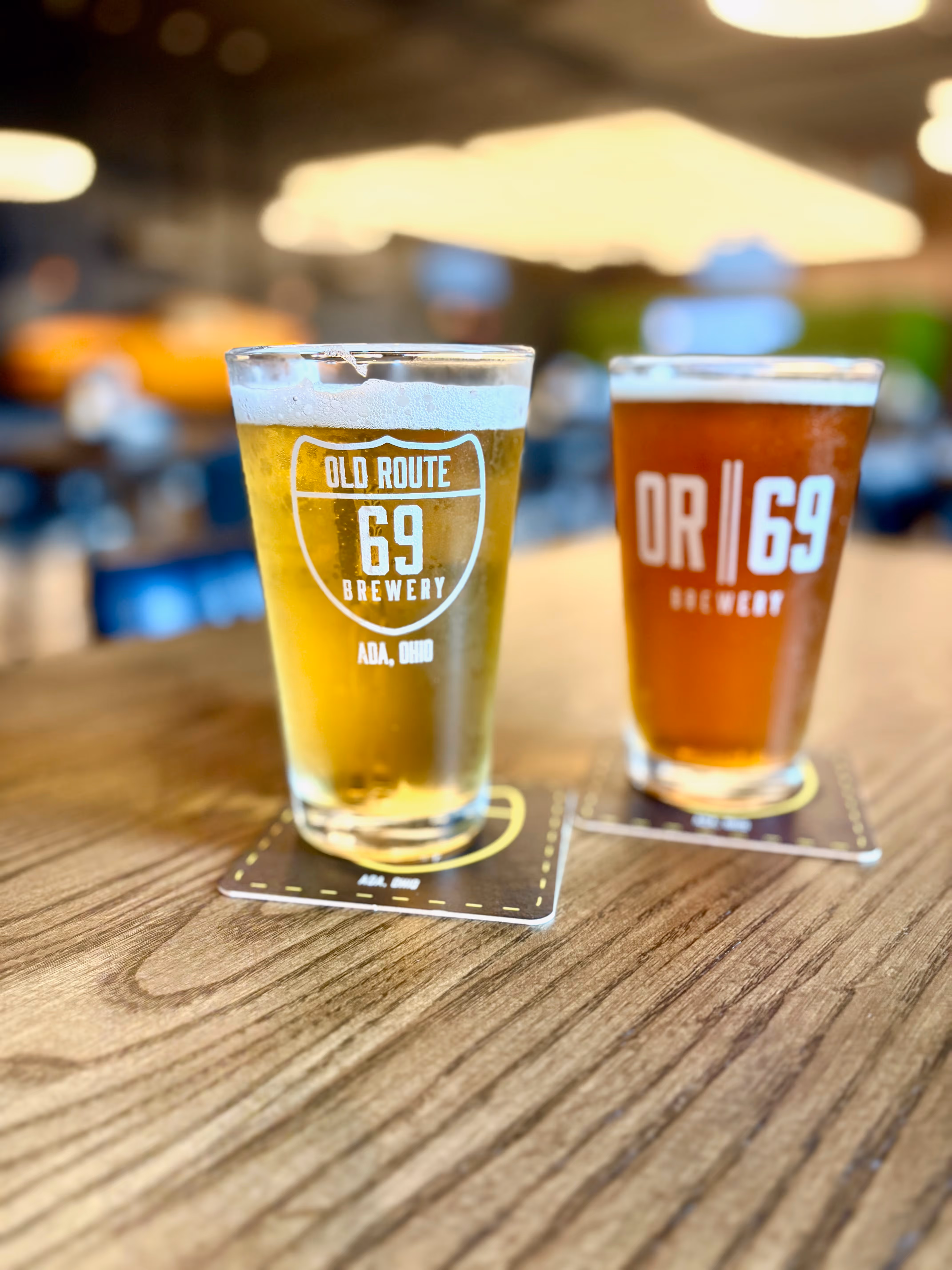 Two pints of craft beer from Old Route 69 Brewery in Ada, Ohio, served on branded coasters atop a wooden table inside the brewery’s taproom.