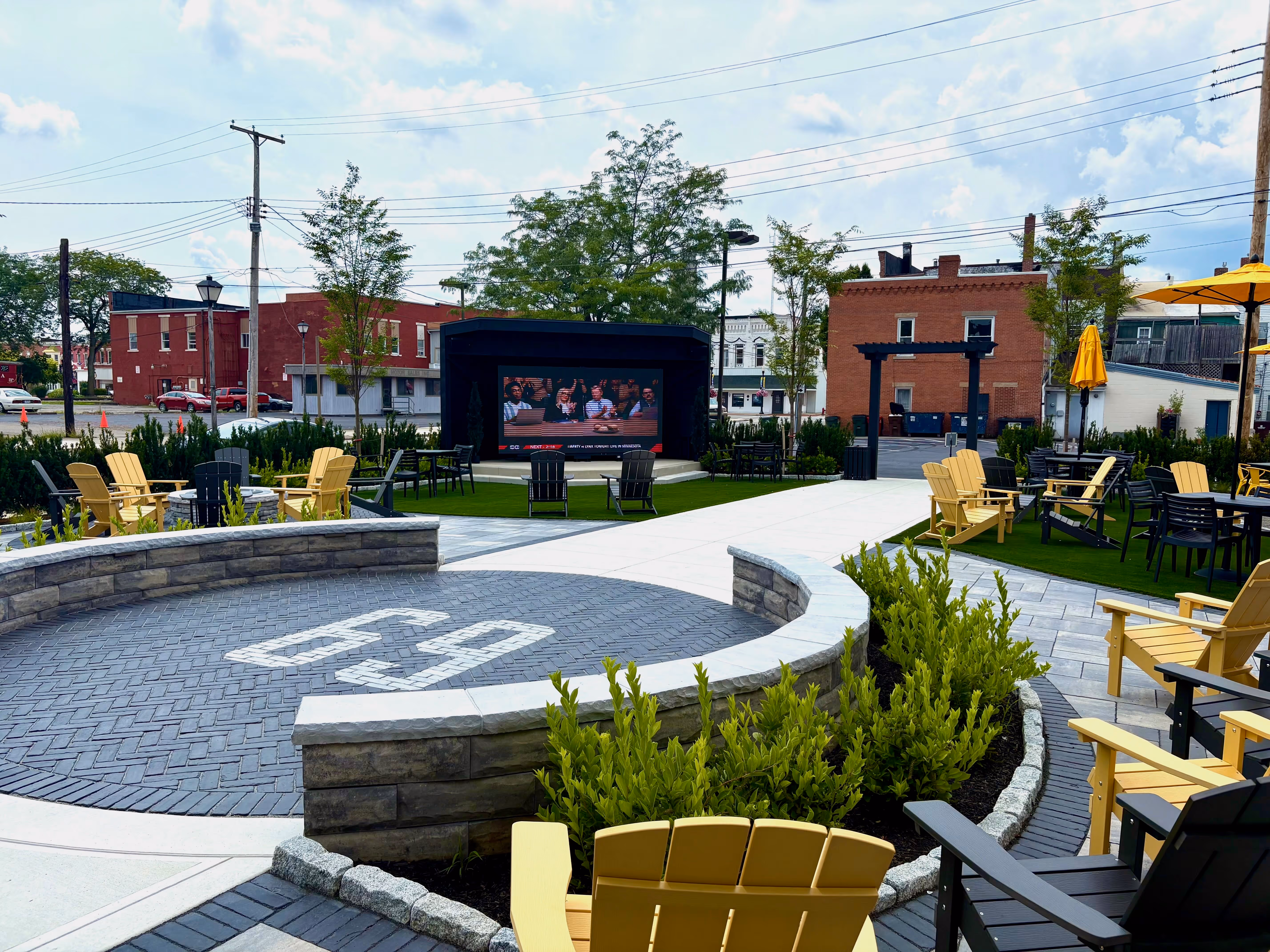 Outdoor beer garden at Old Route 69 Brewery featuring a large central fire pit, lawn seating with Adirondack chairs, and a big screen TV for sports viewing in a vibrant downtown setting.