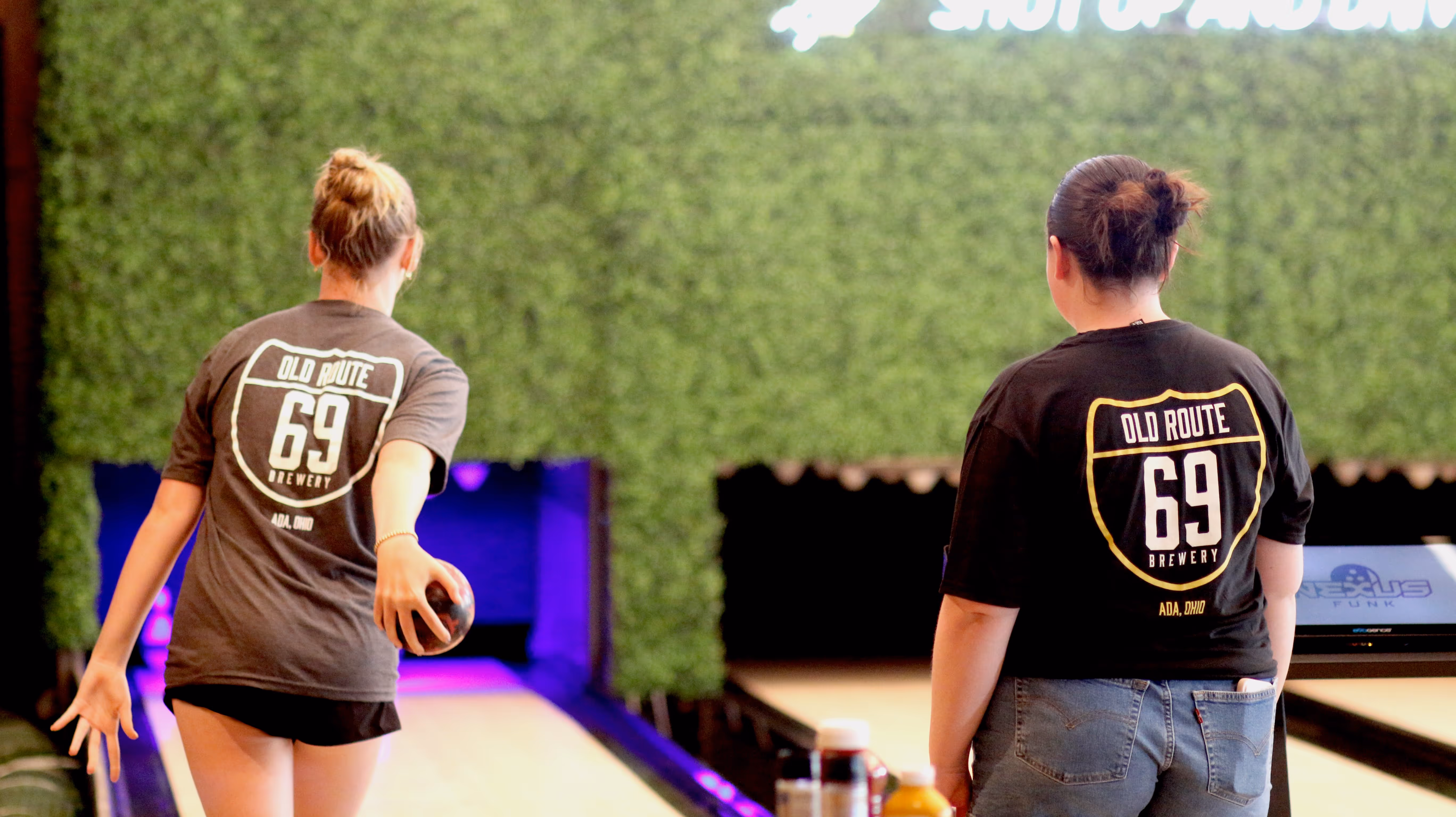 Two women enjoy duckpin bowling at Old Route 69 Brewery in Ada, Ohio, wearing branded brewery shirts. The brewery offers craft beer, entertainment, and unique activities like bowling for private events and group gatherings.