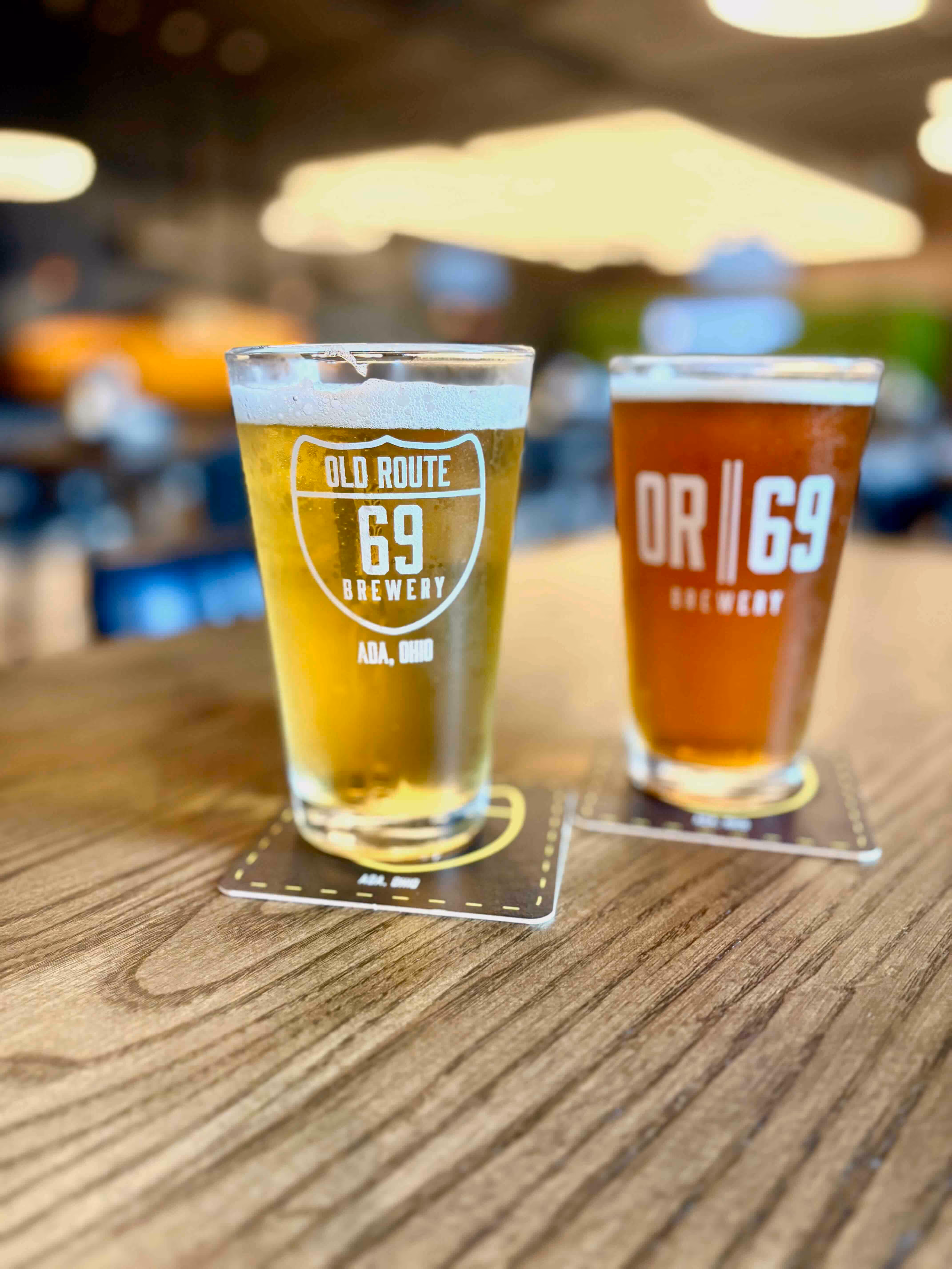 Two pints of craft beer from Old Route 69 Brewery in Ada, Ohio, served on branded coasters atop a wooden table inside the brewery’s taproom.