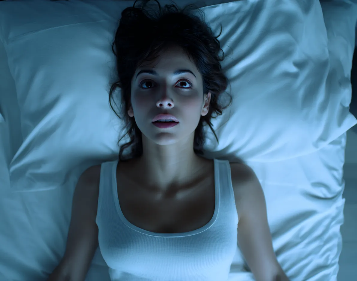 Mujer mexicana de 25 años recostada boca arriba en cama moderna, vista desde arriba, usando camiseta blanca sin mangas, con ojos abiertos y expresión de ligera tensión, iluminada por luz azul tenue nocturna.