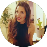 Smiling young woman with long brown hair wearing a black sleeveless top indoors near plants.