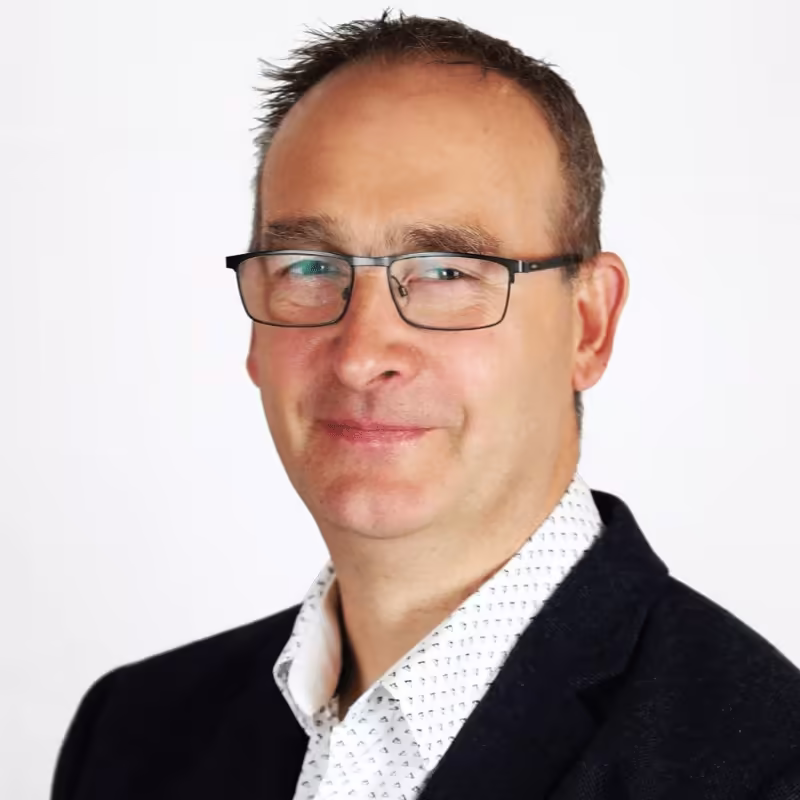 Middle-aged man wearing glasses, a white patterned shirt, and a dark blazer against a white background.