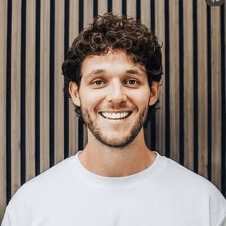Smiling man with curly hair and beard wearing a white shirt in front of vertical wooden panels.
