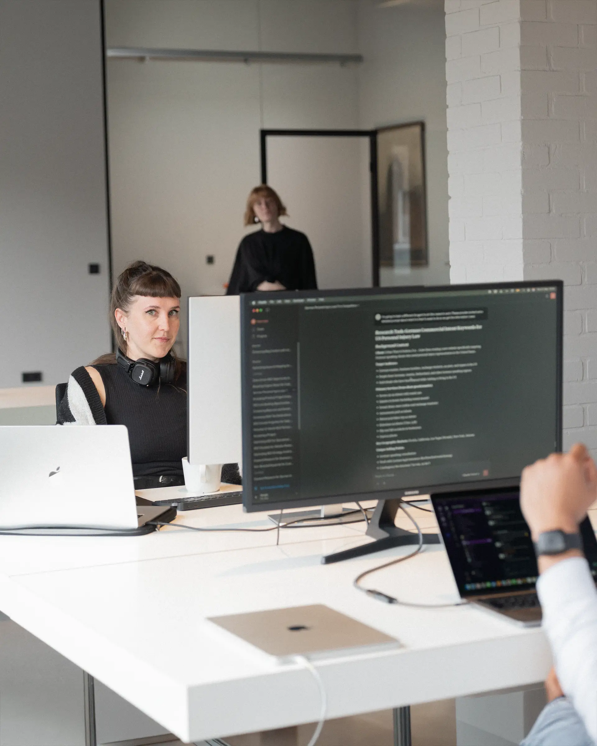 Eine Szene im Büro mit Sergio und Melanie an ihren Plätzen im Vordergrund und vorbeilaufenden Personen im Hintergrund.