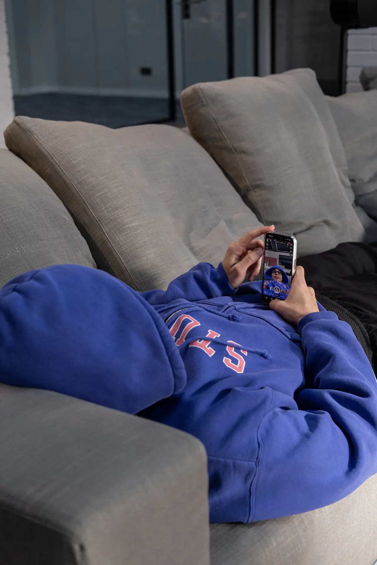 Someone lying on a couch in a blue hoodie watching a video on a phone.