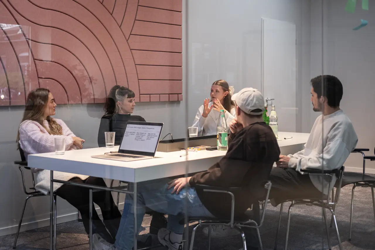 A meeting table scene with four people discussing work around laptops and papers.